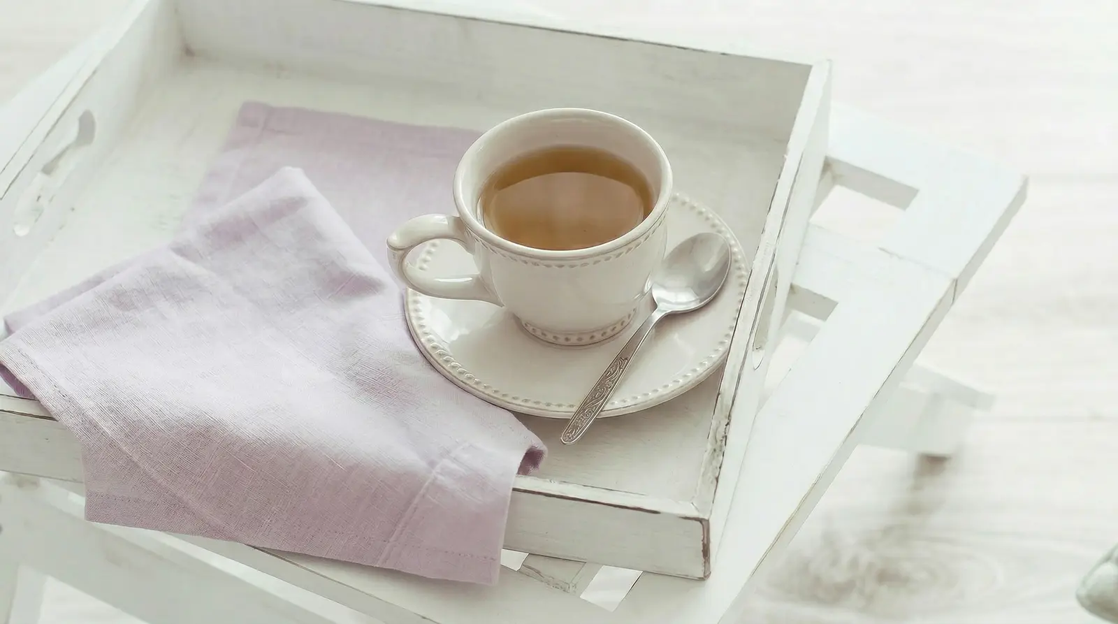 A cup of tea on a white wooden tray with a folded lilac napkin and a spoon.