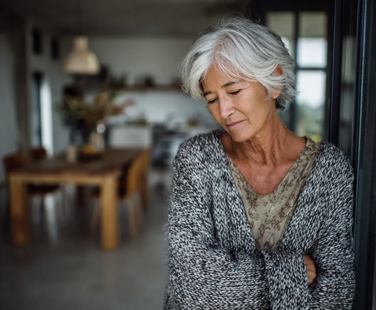 A grey-haired woman standing in a doorway with her arms folded and her eyes lowered.