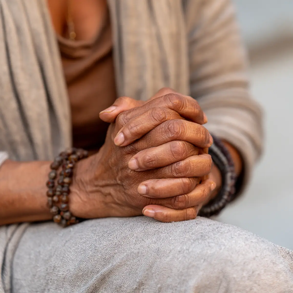 An older woman's hands folded in prayer in her lap, a beaded bracelet on her wrist.
