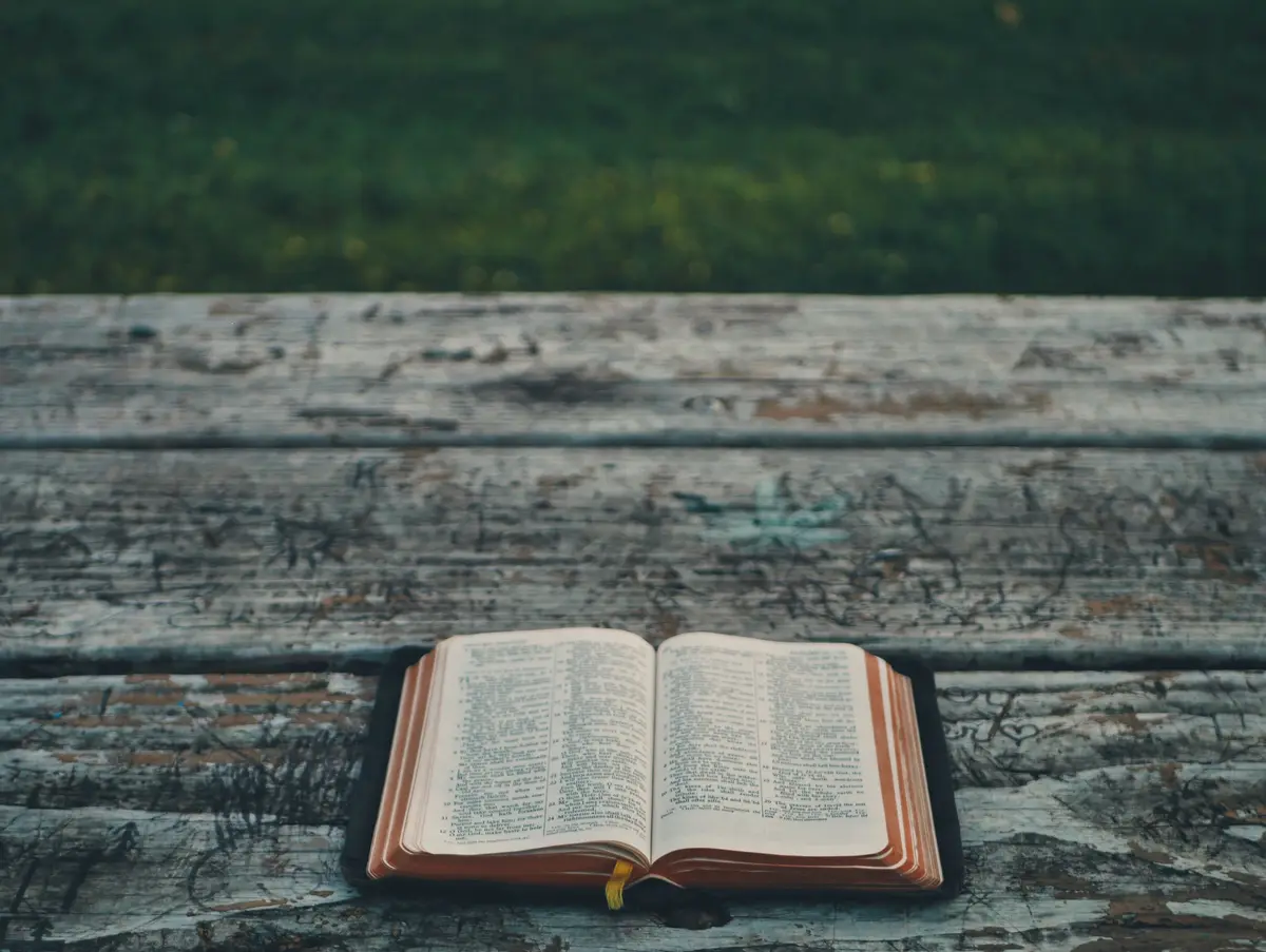 An open Bible lying on a weathered wooden bench outdoors.