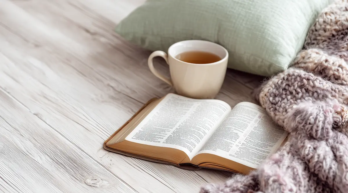 An open Bible on pale wood beside a mug of tea, a sage pillow and a knit throw.